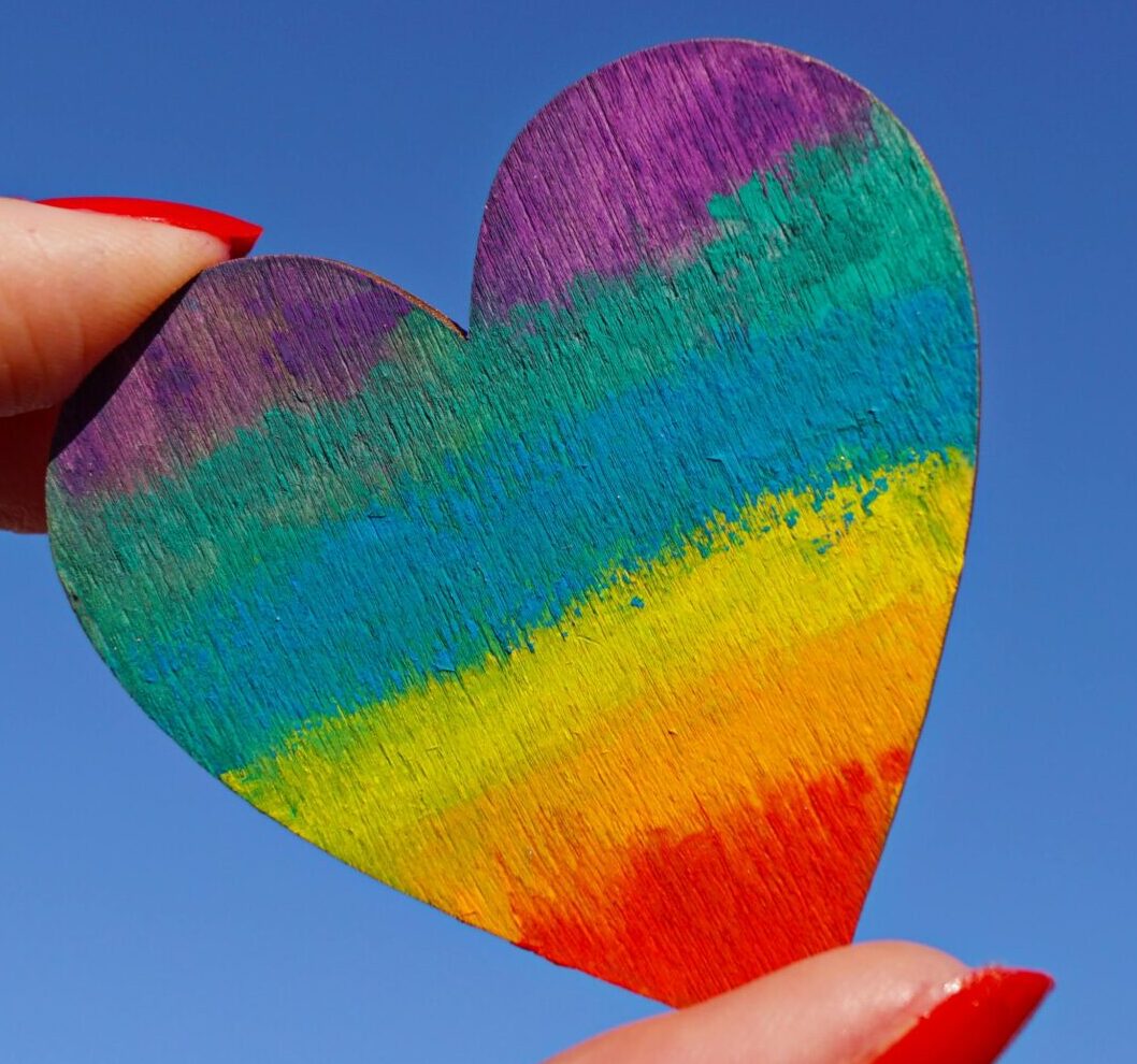 A hand holding a rainbow-colored heart against a clear blue sky, symbolizing love and pride.